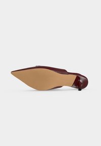 Une seule chaussure à talon haut en cuir bordeaux à bout pointu vue par la semelle montrant une semelle de couleur fauve et un bout de talon noir et élancé.