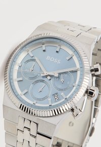 Ασημένιο μεταλλικό ρολόι χειρός με ανοιχτό μπλε καντράν, τρεις υποκαντράν, ένδειξη ημερομηνίας που δείχνει 22 και επωνυμία "BOSS" στο καντράν.