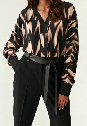 Kobieta w czarno-beżowej bluzce w abstrakcyjne wzory, włożonej do czarnych spodni z wysokim stanem, przewiązanych skórzanym paskiem.