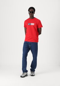 Rød bomulds t-shirt med "Tommy Jeans" logo, blå denim jeans og grå træningssko. Model iført sorte solbriller, stående foran en hvid baggrund.