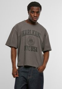 Pelēka, pārmērīga T-kreklā ar melnu apdruku "KARLKANI NYC USA" uz priekšpuses, izgatavota no mīksta kokvilnas auduma. Ikdienišķs dizains ar īsām piedurknēm.