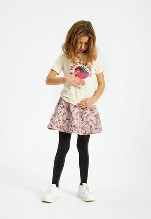Tjej i blommig kjol och svarta leggings som rör vid sitt bröst och mage, och tittar ner på sina händer mot en vit bakgrund.