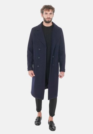 Cappotto doppiopetto in lana navy con ampi revers, dotato di due tasche laterali e lunghezza al ginocchio, indossato sopra un top nero e pantaloni aderenti.