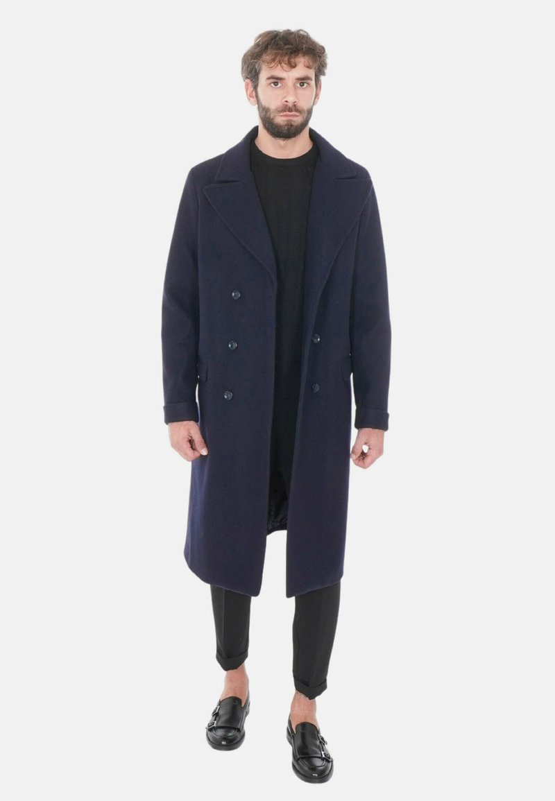 Cappotto doppiopetto in lana navy con ampi revers, dotato di due tasche laterali e lunghezza al ginocchio, indossato sopra un top nero e pantaloni aderenti.