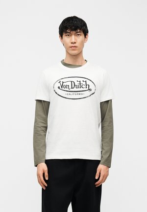 Fiatal férfi, fehér Von Dutch California logós pólót visel zöld hosszú ujjú ing felett, egyszerű háttér előtt áll.