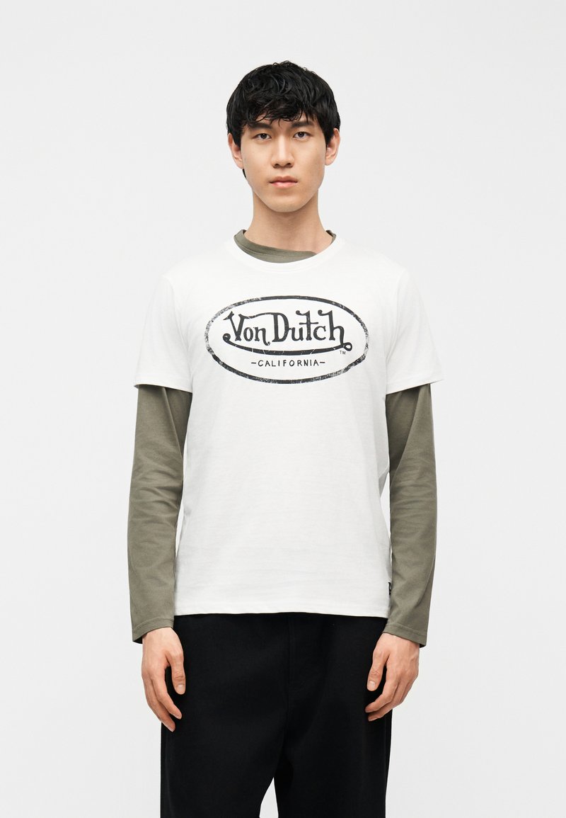 Νεαρός άνδρας που φοράει λευκό T-shirt με το λογότυπο Von Dutch California πάνω από πράσινο πουκάμισο με μακριά μανίκια, στέκεται μπροστά σε απλό φόντο.