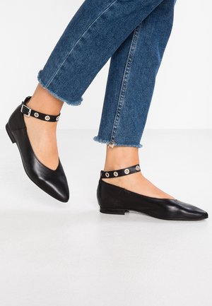 Pies con zapatillas planas negras puntiagudas con tiras en el tobillo con ojales plateados, combinadas con vaqueros azules con dobladillo desgastado sobre un fondo blanco.