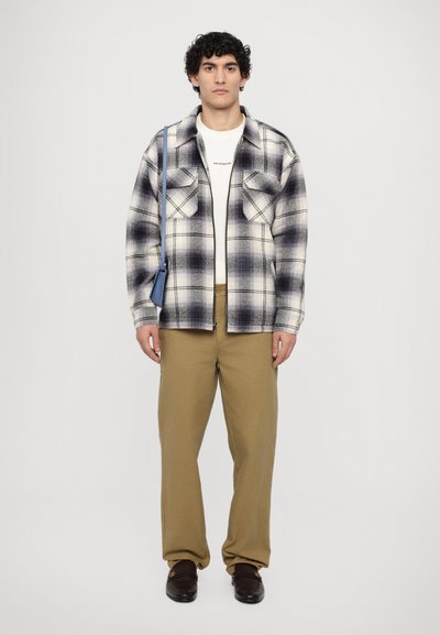 Camisa a cuadros en blanco y morado, con dos bolsillos en el pecho, llevada sobre una camiseta blanca. Pantalones color beige y zapatos marrones sin cordones completan el atuendo.