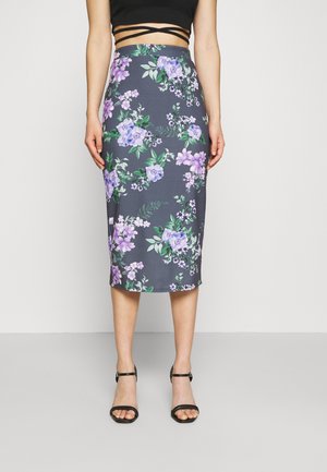 Femme portant une jupe crayon taille haute, de longueur genou, avec un imprimé floral violet et des sandales à talons noires à lanières, sur un fond uni.