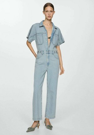 Denim jumpsuit i lyseblå, korte ærmer, knaplukning foran, bæltet talje, bred ben design. Båret med grå højhælede sko.