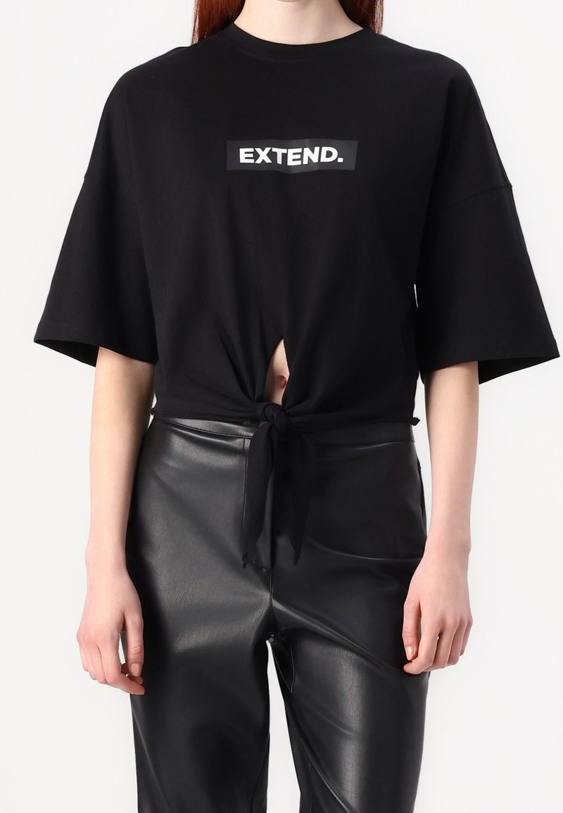T-shirt noir à manches courtes avec un détail nœud à l'avant, arborant un graphique blanc "EXTEND." sur la poitrine. Associé à un pantalon noir brillant.