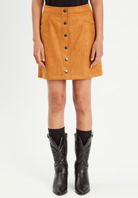 Mini-jupe en suède beige avec une fermeture à boutons sur le devant, deux poches latérales et une coupe droite. Associée à des bottes de cowboy noires et des chaussettes sombres.