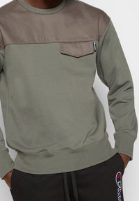 Uomo che indossa un felpa bicolore verde oliva e marrone con taschino e pantaloni della tuta neri con patch del logo Champion.