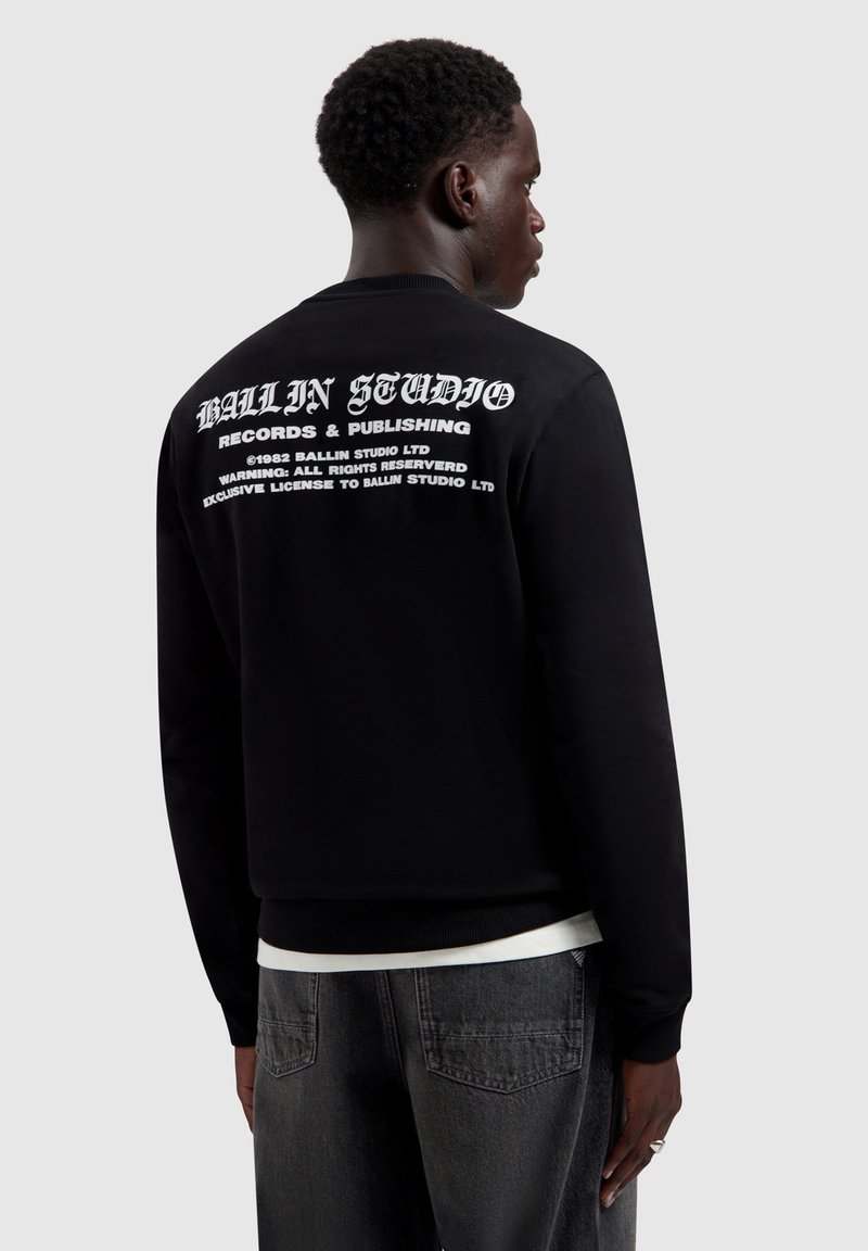 Man in een zwart sweatshirt met witte tekst "BALLIN STUDIO Records & Publishing" op de achterkant, gecombineerd met een donker spijkerbroek, kijkend naar de zijkant.