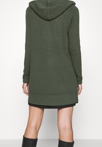 Dlouhá olivově zelená pletená mikina s kapucí, texturovaným vzorem, přiléhavými rukávy a rovný střihem. Má kapuci a žebrovaný lem.