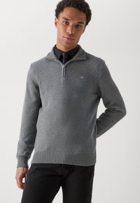 Grauer Strickpullover mit hohem Kragen und Reißverschluss. Mit gerippten Bündchen und Saum. Über einem schwarzen Shirt getragen. Einfaches Logo auf der Brust.