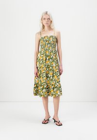Robe midi fleuris verts avec des fruits jaunes, corsage smocké, fines bretelles et jupe à volants. Portée avec des sandales à talons noires.