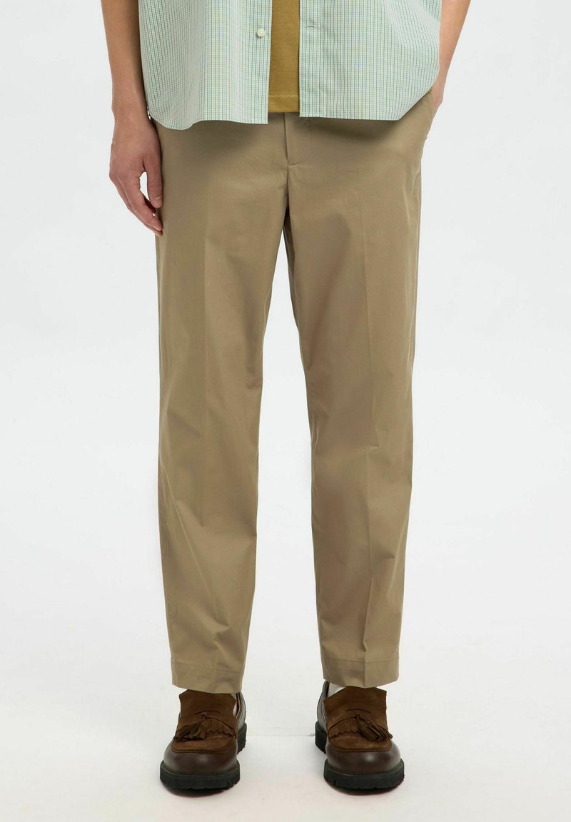 Beige rechte broek van gladde stof, met een strakke uitstraling en subtiele plooien, gecombineerd met bruine schoenen.