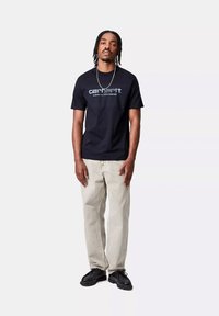 T-shirt nera in cotone con loghi bianchi "carhartt WORK IN PROGRESS". Abbinata a jeans grigi chiari e scarpe nere, in piedi contro uno sfondo semplice.