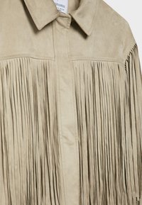 Veste en daim beige avec de longues franges verticales sous la poitrine et sur les manches, avec un col plié.