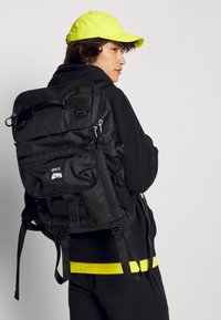 Czarny plecak z wieloma przegródkami i regulowanymi paskami, w połączeniu z neonowo-żółtą czapką, charakteryzujący się eleganckim materiałem i wzornictwem.