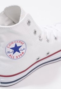 Bílé vysoké tenisky Converse Chuck Taylor All Star s červenými a modrými pruhy na podrážce a podpisovou kulatou nášivkou na boku.