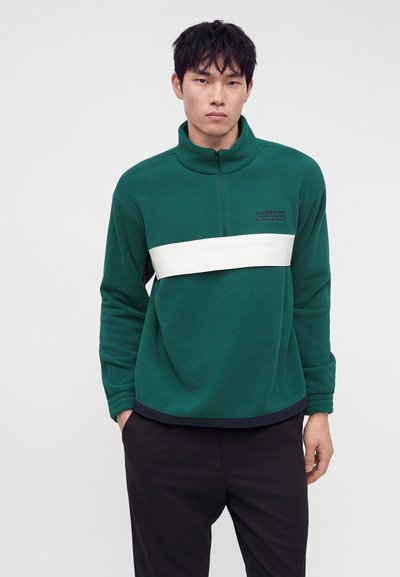 Νέος άνδρας που φοράει πράσινο fleece με φερμουάρ στο ένα τέταρτο, με λευκή οριζόντια λωρίδα και μαύρο παντελόνι, στέκεται μπροστά σε απλό φωτεινό φόντο.