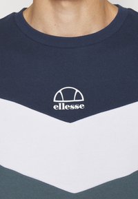 Sudadera de cuello redondo en azul marino, blanco y verde azulado con un diseño de chevron. Presenta el logo "ellesse" en blanco en el pecho. Material suave de algodón.