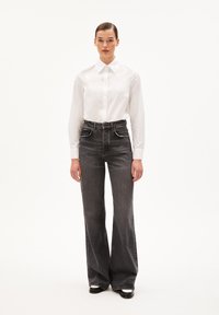 Camicia bianca con colletto classico, abbinata a jeans neri a vita alta svasati. Tessuto liscio e vestibilità sartoriale. Scarpe nere.