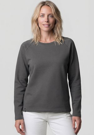 Blonde Frau trägt ein dunkelgraues Langarmshirt und weiße Hose, steht und schaut nach vorne vor neutralem Hintergrund.