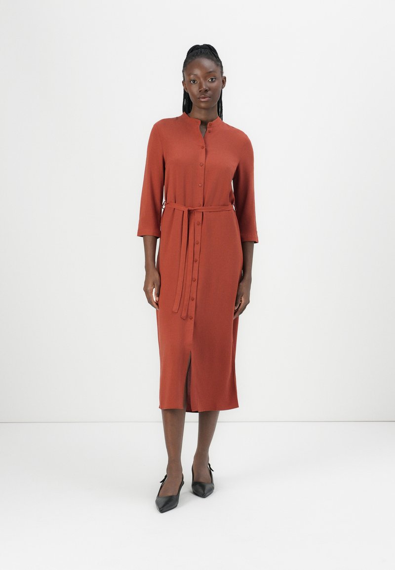 Robe mi-longue de couleur rouille avec un devant à boutons, des manches trois-quarts, une ceinture à la taille et une fente à l'avant. Tissu lisse.