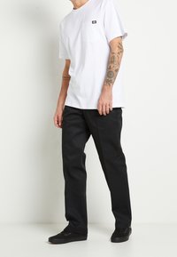 Camiseta de algodón blanca con bolsillo y parche de logo, combinada con pantalones negros de pierna recta y zapatillas negras, mostrando un atuendo informal.