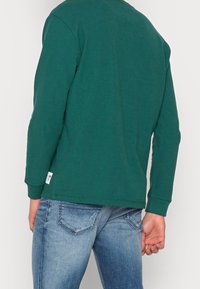 Zelená košile s dlouhým rukávem vyrobená z bavlny. Má kulatý výstřih, ribované manžety a štítek na spodním lemu. V kombinaci se světle modrými džínami.