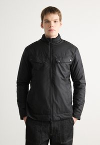 Schwarze Zip-Jacke mit hohem Kragen, zwei Brusttaschen mit Klappen und glatter Textur. Getragen über dunklen Jeanshosen.