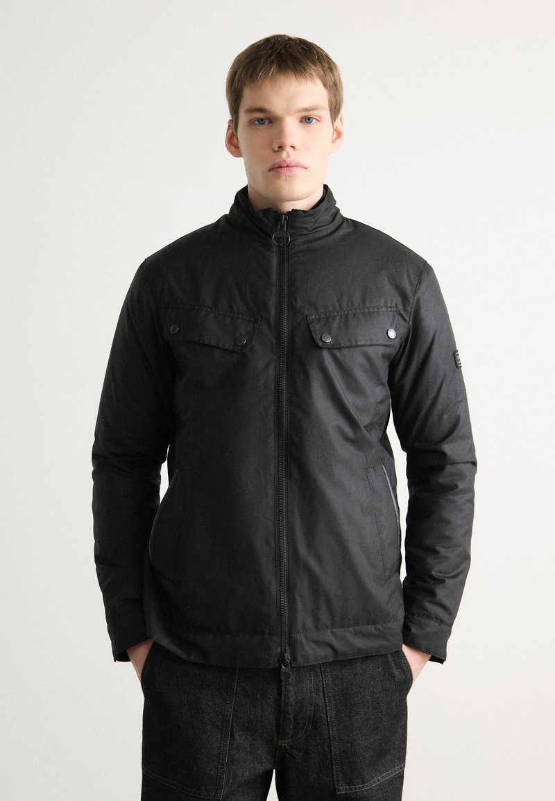 Schwarze Zip-Jacke mit hohem Kragen, zwei Brusttaschen mit Klappen und glatter Textur. Getragen über dunklen Jeanshosen.