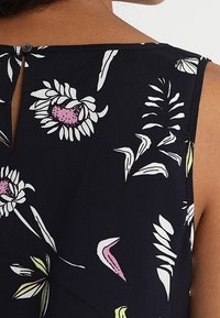 Granatowa bluzka w kwiaty z białymi, różowymi i zielonymi wzorami, z otworem w kształcie łezki z tyłu oraz pojedynczym, okrągłym guzikiem przy dekolcie.