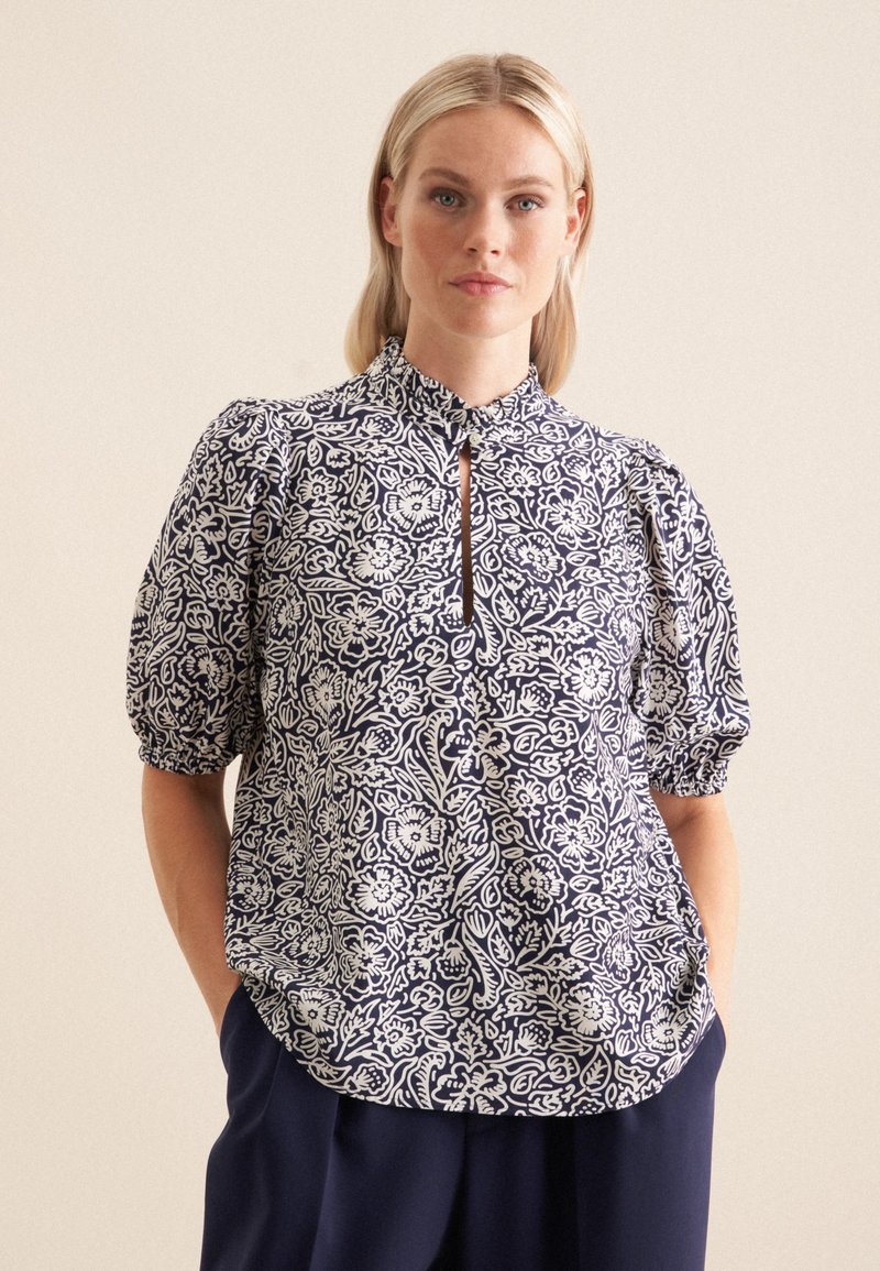 Blusa blu navy con motivo floreale bianco, maniche a sbuffo corte, scollo a chiave e tessuto morbido. Presenta una vestibilità rilassata e dettagli arricciati.