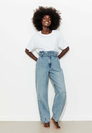 Femme souriante, pieds nus, avec une grande afro, porte un t-shirt blanc rentré dans un jean bleu taille haute ceinturé, debout contre un mur blanc.