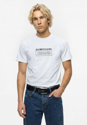 Blond muž nosiaci biele tričko Karl Lagerfeld Jeans zastrčené do modrých džínsov s čiernym opaskom, stojaci na bielom pozadí.