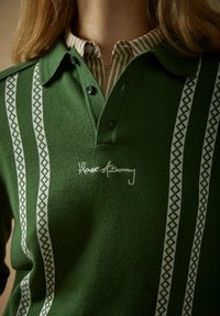 Roheline kootud polosärk valgete mustriliste vertikaalsete triipudega ja „House of Sunny” tikandiga rinnal, kandmas triibulise nööpidega särgi kraega.