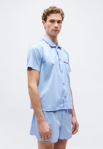Νέος άνδρας που φοράει ελαφρύ γαλάζιο πουκάμισο με κοντά μανίκια και matching σορτς, στέκεται σε απλό λευκό φόντο.