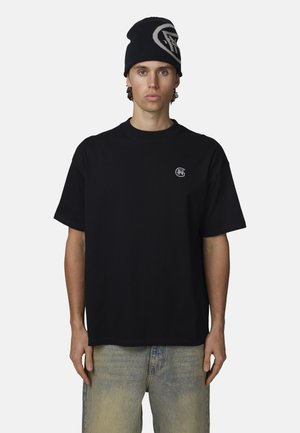 Giovane uomo con berretto nero con logo, t-shirt nera oversize con piccolo logo sul petto e jeans blu sbiaditi, in piedi su sfondo bianco.