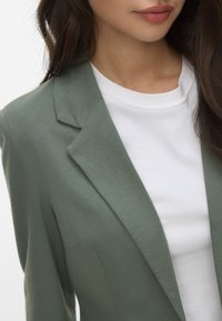 Blazer vert avec un revers de col, associé à un t-shirt blanc uni. Le tissu semble doux et lisse avec une silhouette ajustée.