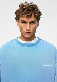 Lichtblauwe sweatshirt met ronde hals en ribbelkraag. Voorzien van witte tekst "ESSENCE PROJECT" aan de voorkant. Zachte, gestructureerde stof.