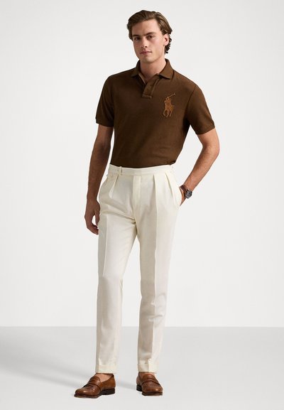 Cămașă polo din bumbac maro, cu logo brodat, asociată cu pantaloni albi cu pliuri. Modelul poartă mocasini din piele maro și un ceas.