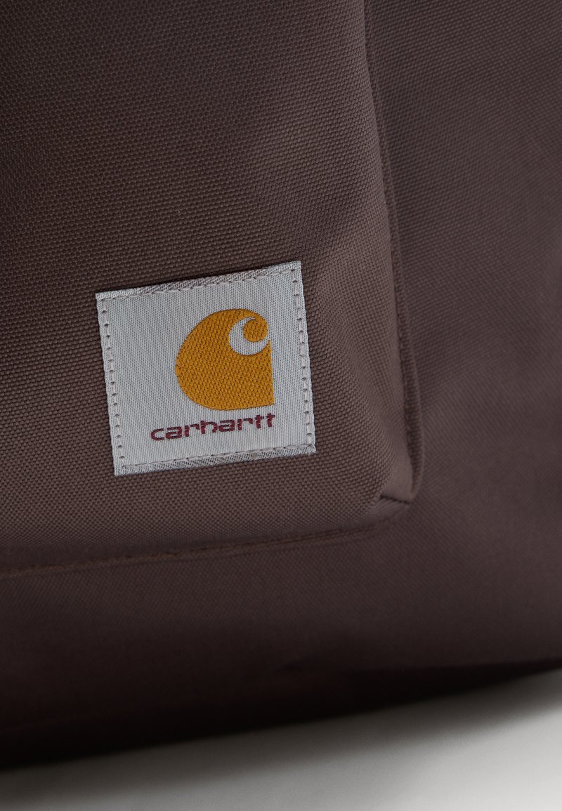 Tissu marron avec une étiquette carrée blanche comportant un logo Carhartt jaune cousu sur la surface.