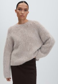 Woman with dark hair in a low bun wearing a loose, fuzzy light beige sweater and a dark skirt against a plain light gray background.