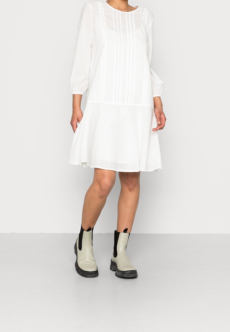 Femme portant une robe blanche à manches longues, longueur genou, avec des rayures verticales, accompagnée de bottines grises claires avec des côtés élastiques noirs.