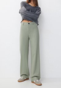 Pantalons à jambes larges en tissu vert clair à la texture lisse, dotés d'une fermeture à bouton à l'avant et de poches, associés à un haut en maille grise.
