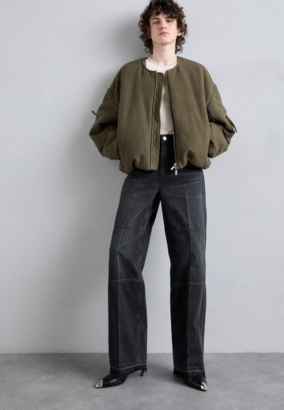 Veste bomber vert olive avec col côtelé, jean noir taille haute à jambes larges avec détails de couture, et escarpins noirs à bout pointu avec accents en argent.